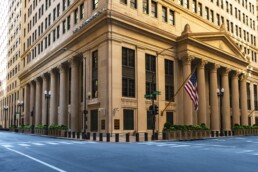 Large tan building with windows and pillars around it, an American flag hanging at the front and a Federal Reserve Bank sign at the corner