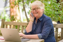 Senior woman sitting on her porch, talking on her phone and using a laptop to review her tax-efficient retirement savings