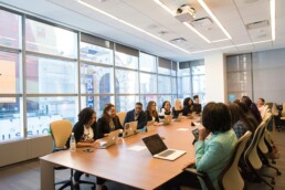 a large group of investors sat around a wooden meeting table in an office with windows, discussing ethical investment opportunities