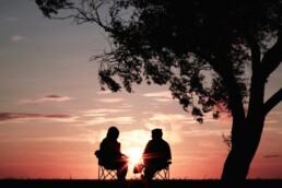 Silhouettes of two seniors enjoying their retirement savings, sitting under a tree in lawn chairs and watching the sunset