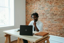 A well-dressed man sitting at a desk in front of an exposed brick wall, exploring emerging markets on his laptop PC