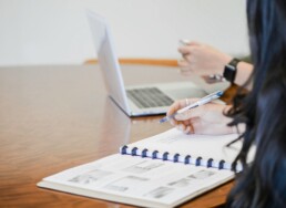 One person sitting at a desk and using a pen and ledger to calculate compound interest while a second person uses a laptop and smartphone in the background