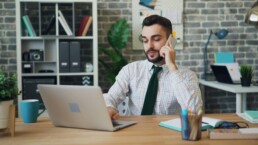 A professionally-dressed financial advisor sitting at a desk, using a silver laptop and talking to a client on a smartphone