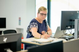 A blond middle-aged woman sitting at a desk and using a laptop to monitor her passive income so that she can take notes in a ledger