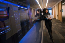 A young woman who is investing in technology standing in a corporate hallway, reviewing information on her laptop while standing near a room full of computer hardware