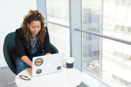 A young woman sitting at her desk with a cup of coffee by a large window, exploring entrepreneurial finance on her computer to grow her business