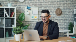 A man in business attire sitting at a desk in front of an exposed brick wall, performing stock market analysis on his silver laptop