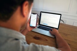 A man sat at a desk, comparing finance charts on his smartphone and laptop to determine how his index funds are performing.