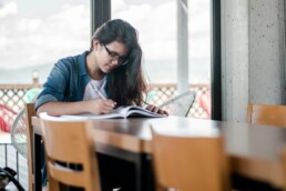 A young woman sitting at a table in front of a sliding glass door, planning out her budget to avoid making common financial mistakes.