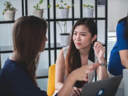 Two young women sitting at a desk in an office setting, discussing financial boundaries